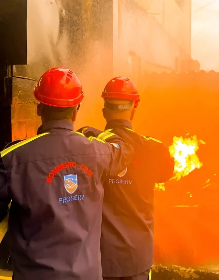 Bombeiros combatendo princípio de incêndio em container com extintor de espuma em área externa com vegetação e piso de paralelepípedos.