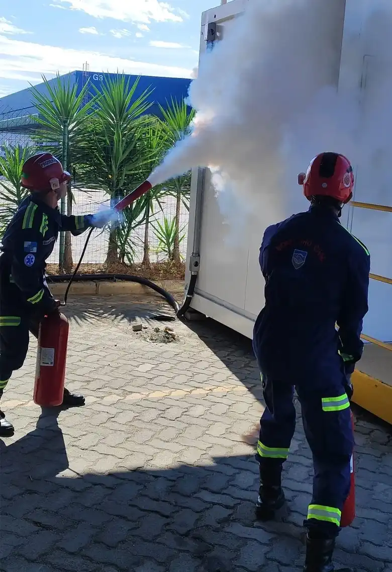 Bombeiros atuando no combate a incêndio em construção, utilizando equipamentos de proteção individual com chamas visíveis ao fundo.